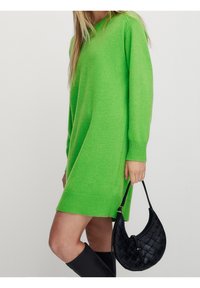 Robe tricotée verte à manches longues, poignets et ourlet côtelés. Associée à un sac à main tissé noir et des bottes noires montant jusqu'aux genoux.