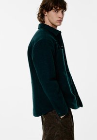 Giacca in pile verde con colletto, bottoni sul davanti e tasca laterale. Tessuto strutturato e vestibilità comoda, abbinata a pantaloni marroni con fantasia.