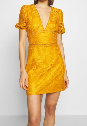 Femme portant une courte robe en dentelle jaune moutarde avec des manches courtes bouffantes et un décolleté en V profond.