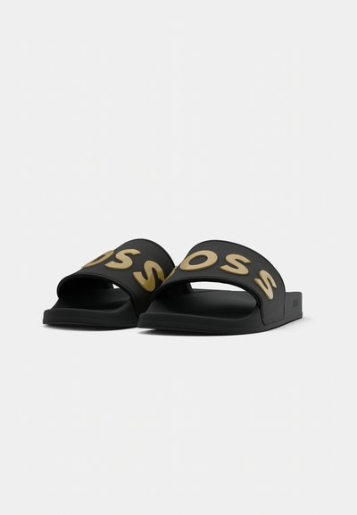 Tongs noires avec des larges brides affichant des lettres en or, présentant une finition mate lisse et un design de semelle intérieure ergonomique.