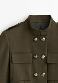 Veste vert olive avec un col haut, design à double boutonnage et boutons en laiton. Dispose de deux poches avant et d'une texture lisse.