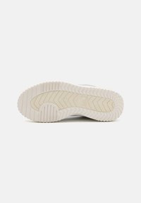 White sneaker outsole with a textured rubber surface, featuring a zigzag tread pattern and embossed logo detail near the arch.