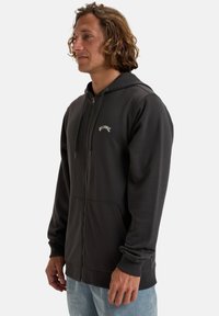 Sort zip-up hoodie med en kængurulomme, justerbar hætte med snøre og broderet logo på brystet. Tætsiddende manchetter og blød tekstur.