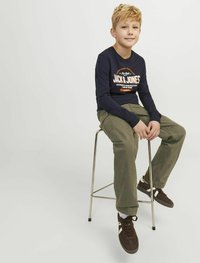 Mornarica dolgi rokavi srajca z logotipom "Jack & Jones", olivno zeleni hlače, rjavi superge, sedeč na kovinskem stolčku ob enobarvni podlagi.