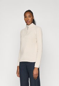 Pull en beige clair, texturé, avec un col zippé quart et des manches longues, associé à un pantalon foncé. Design simple et minimaliste.