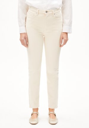 Femme portant un jean slim blanc cassé, une chemise blanche à manches longues boutonnée, et des chaussures style Mary Jane beige clair, debout sur fond blanc.