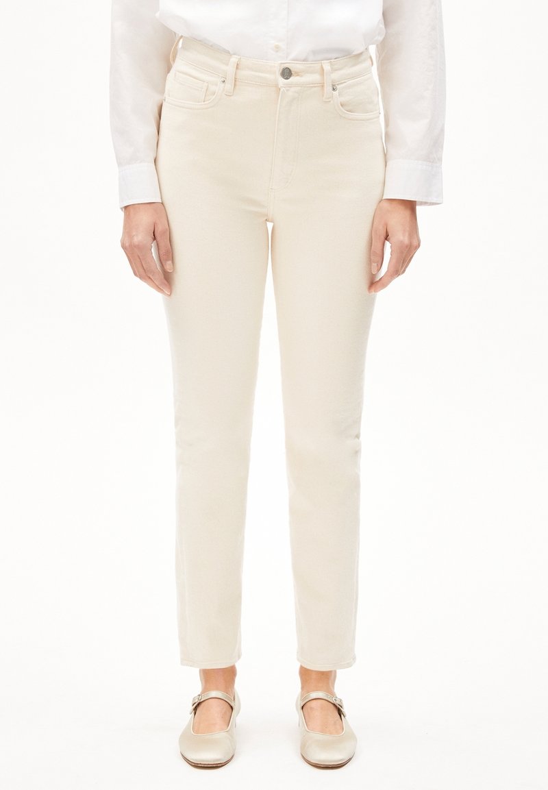Femme portant un jean slim blanc cassé, une chemise blanche à manches longues boutonnée, et des chaussures style Mary Jane beige clair, debout sur fond blanc.