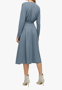 Kobieta w niebieskiej midi sukience z długim rękawem, marszczonym pasem, z guzikami na mankietach i kremowymi botkami, stoi plecami na białym tle.