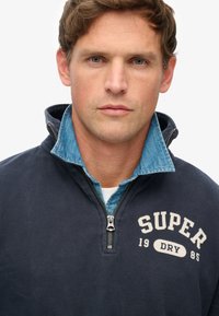 Mann mit hellbraunen Haaren und blauen Augen, der einen marineblauen Sweatshirt mit Viertelreißverschluss trägt, auf dem "SUPER DRY 1985" steht, über einem Denim-Hemd mit Kragen.