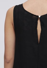 Top senza maniche nero con design a chiave sulla schiena, caratterizzato da una chiusura con piccolo bottone rotondo. Il materiale sembra leggero e liscio.
