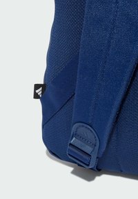 Niebieski plecak z materiału o teksturowanej powierzchni, z wyściełanymi paskami i plastikowym zapięciem, z małą metką z logo po boku.