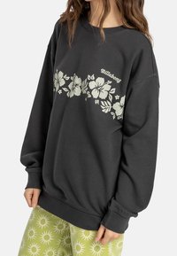 Sudadera negra de gran tamaño con estampado floral blanco y texto "Billabong", hecha de una tela suave, con mangas largas y puños de canalé.