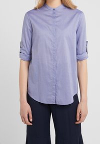 Camicia azzurra chiara, con bottoni e colletto alla coreana, maniche arrotolabili e orlo leggermente curvo. Realizzata in tessuto intrecciato con una texture liscia.