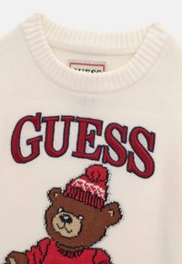 Pull blanc en maille avec un ours brun portant un bonnet d'hiver rouge et un pull, et le mot GUESS en grandes lettres rouges au-dessus.
