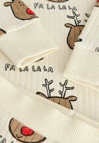 Kremowa ribbed tkanina z kreskówkowymi twarzami reniferów z czerwonymi nosami oraz napisem "FA LA LA LA" w kolorze czarnym. Miękka tekstura i prosty, świąteczny wzór.