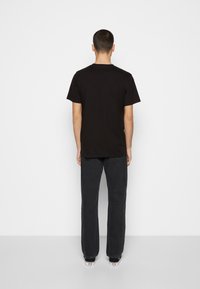 Mann steht mit dem Gesicht zu einer weißen Wand, trägt ein schwarzes T-Shirt, dunkle Jeans und schwarze Sneakers mit weißen Sohlen.