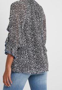 Genomskinlig svart blus med ett vitt blommönster, volangärmar med blå accenter och en avslappnad passform. Styla gärna med ljusblå jeans.