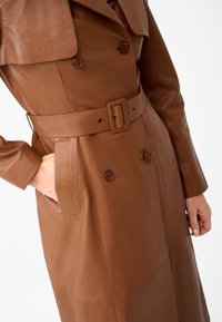 Brown leather trench coat featuring a double-breasted design, wide lapels, a belted waist, and side pockets. Smooth texture with button details.
