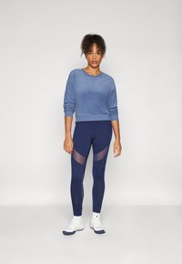 Donna in felpa blu, leggings navy con pannelli a rete, calzini bianchi e scarpe da ginnastica bianche su uno sfondo chiaro e neutro.