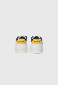 Des baskets blanches avec des accents verts et jaunes, présentant une texture lisse, une semelle plate et un logo sur le talon. Vue arrière affichée.