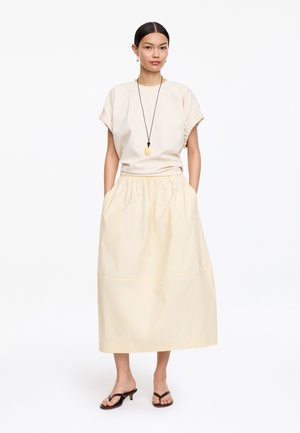 Mujer con blusa beige de manga corta y falda larga, de pie con las manos en los bolsillos de la falda, complementada con un collar largo colgante y sandalias de tacón.