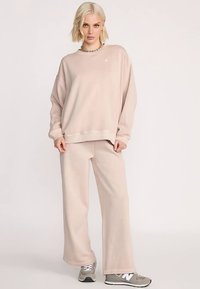 Felpa in pile beige con taglio comodo e collo a giro abbinata a pantaloni a gamba larga coordinati. Le sneakers presentano dettagli grigi e bianchi.