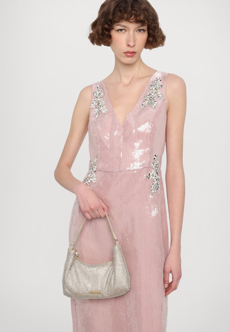 Roze paillettenjurk met v-hals, versierd met zilveren bloemapplicaties. Model met een metalen zilveren hobo tas. Eenvoudige achtergrond.