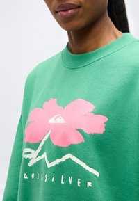 Κοντινό πλάνο ενός ατόμου που φοράει ένα πράσινο φούτερ με ένα μεγάλο ροζ λουλούδι και λευκές αφηρημένες γραμμές και κείμενο στο μπροστινό μέρος.