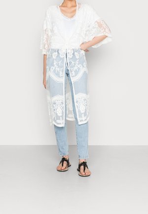 Femme portant un jean bleu clair, des sandales noires, un débardeur blanc et un long cardigan en dentelle blanche noué à la taille, debout avec une main sur la hanche.