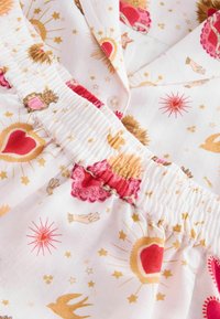 Tela blanca con estallidos de estrellas doradas, soles, corazones rojos, corazones coronados rosas, pequeñas manos beige y estampados de pájaros dorados, con cintura elástica.