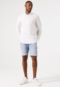 Witte linnen blouse met lange mouwen, lichtblauwe opgerolde denimshorts, witte sneakers, met zichtbare been tattoo; minimalistisch ontwerp, casual fit.