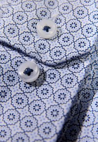 Tessuto bianco con motivo floreale blu, con due bottoni trasparenti cuciti con filo blu. Dettaglio del colletto e dei bordi cuciti visibili.