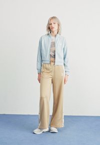 Chaqueta bomber azul clara con puños acanalados, combinada con pantalones anchos beiges con botones de lado. Zapatillas grises completan el atuendo.