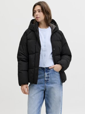 Sieviete melnā pūkainā jakā virs gaiši zilas pogātas kardiganas un brīvām zilām džinsu biksēm, stāv ar vienu roku kabatā.