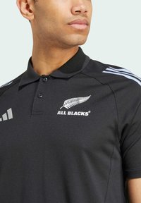 Polo nero realizzato in tessuto testurizzato. Presenta un colletto con bottoni, il logo bianco "ALL BLACKS" e strisce azzurre chiare sulle spalle.