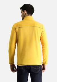 Chaqueta de forro polar amarilla con un cuello alto y mangas largas. Presenta costuras en contraste y una textura suave. Se lleva con jeans oscuros.