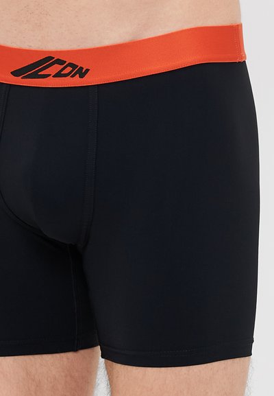 Juodi trumpi šortai su oranžine elastinga juosmeniu, ant kurio yra logotipas, pagaminti iš lygaus audinio. Jie turi prigludusį dizainą ir minimalų siuvimą.