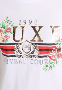 Camiseta blanca con el texto "LUXE" impreso en dorado, acentos florales en rosa y verde, y detalles a rayas en negro y rojo.