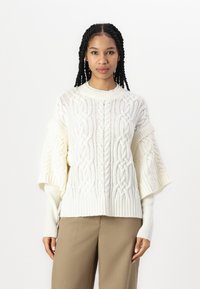 Maglione crema a trecce con maniche larghe, orlo a coste e motivi testurizzati. Abbinato a pantaloni beige, presentato su uno sfondo bianco.