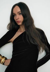 Melna zīda kleita ar asimetrisku griezumu, garām piedurknēm, kas izgatavotas no caurspīdīgas auduma ar punktiņiem, un zeltīta aproce uz plaukstas. Gluda tekstūra, pieguļošs dizains.