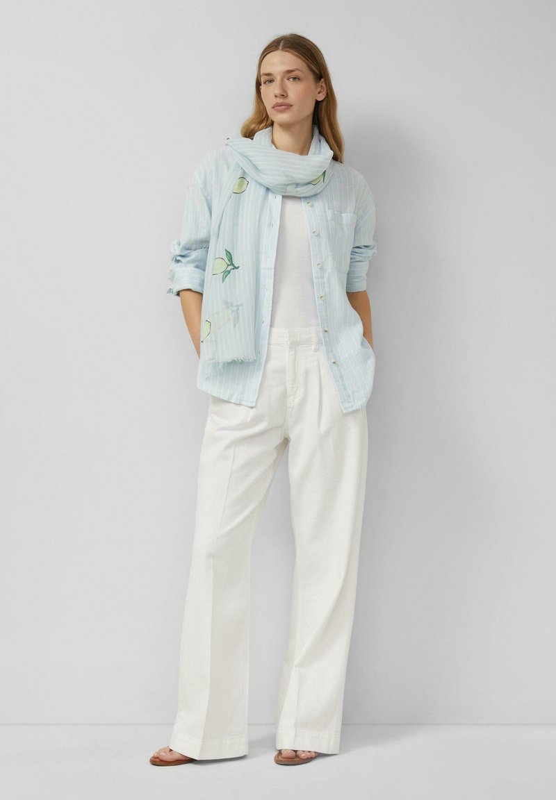Femme debout contre un mur uni portant une chemise rayée bleu clair, un pantalon blanc, une écharpe avec des imprimés citron et des sandales.