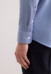 Primo piano della mano sinistra e del polso di una persona che indossa una camicia a maniche lunghe blu a fantasia con bottoni bianchi e pantaloni scuri su uno sfondo chiaro.