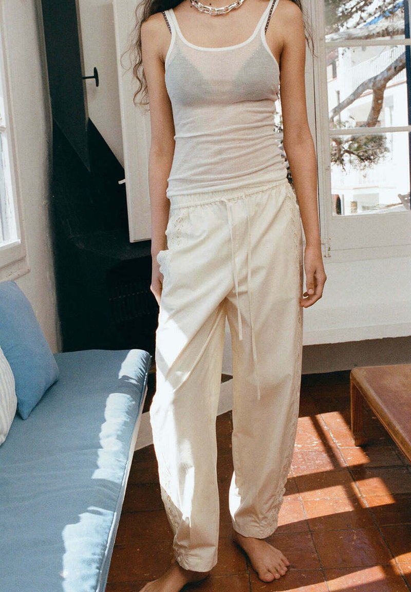 Femme debout pieds nus à l'intérieur, portant un débardeur blanc transparent et un pantalon ample crème avec des côtés brodés, avec la lumière naturelle à travers les fenêtres.
