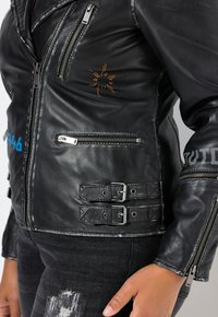 Schwarze Lederjacke mit Reißverschlusstaschen, orangefarbener Stickerei und Schnallenakzenten an der Taille. Silberne Hardware und abgenutzte Textur.