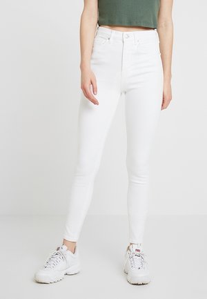 Personne portant un jean skinny blanc taille haute, un top court vert côtelé et des baskets blanches épaisses, debout sur un fond clair uni.