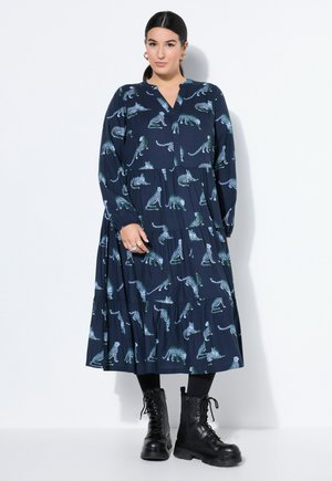 Kvinde iført en navyblå midi kjole med hvide og grønne leopardtryk, sorte leggings, sorte snørebånds combat-støvler og gyldne hoop-øreringe.