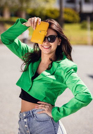 Green cropped jacket made of smooth fabric, paired with a black top and high-waisted denim shorts. Holding a yellow wallet, wearing sunglasses.