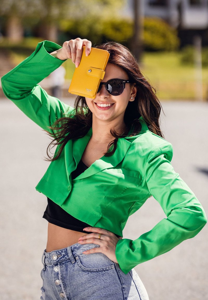 Green cropped jacket made of smooth fabric, paired with a black top and high-waisted denim shorts. Holding a yellow wallet, wearing sunglasses.