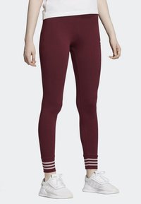 Burgundowe legginsy wykonane z elastycznego materiału, z białymi paskami przy kostkach oraz dopasowanym krojem. Noszone z białymi sneakersami.