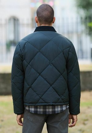 Dark green quilted jacket with a diamond pattern, featuring a black collar and layered over a blue plaid shirt, gray pants visible.